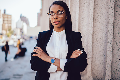 Confident African American female entrepreneur in formal clothing and eyeglasses standing with crossed hands and looking at camera in New York street