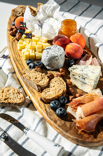 Wooden board with variety of cheeses, prosciutto, bread, blueberries, apricots, jam and nuts