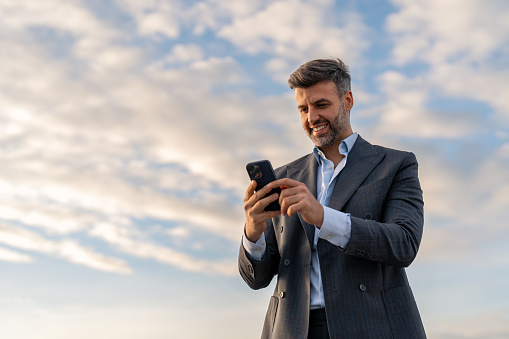 Smiling handsome professional businessman worker, holding smartphone standing outdoors using mobile technology, looking at cell phone, texting on cellphone, sending SMS, typing on cellular technology device, checking corporate apps, browsing online.