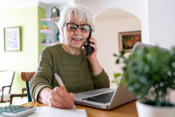 Senior woman working at home and talking on the phone while using her laptop