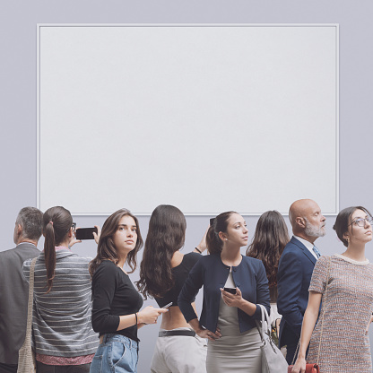 Crowd of visitors at the art gallery, they are looking at paintings and taking pictures, blank painting frame in the background