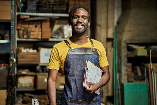 Portrait of a happy young male carpenter wearing apron holding a clipboard with drawing standing in his workshop