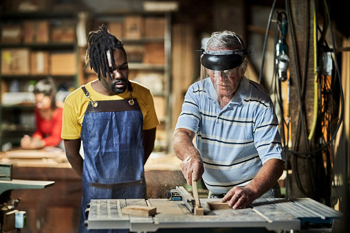 Senior man sawing wooden plank on machine in woodwork class with a carpenter