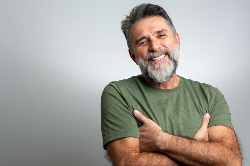 Portrait of attractive mature sporty man in green t-shirt standing with crossed arms isolated on gray background. Happy casual man smiling and holding his hand crossed at his chest, standing on white