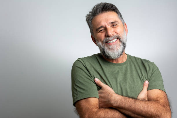 Portrait of a mature guy with arms folded Portrait of attractive mature sporty man in green t-shirt standing with crossed arms isolated on gray background. Happy casual man smiling and holding his hand crossed at his chest, standing on white 55 59 years stock pictures, royalty-free photos & images