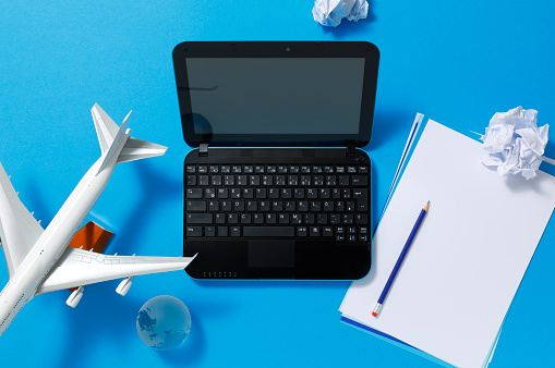 Netbook with blank sheets of paper with a blue pencil and crumpled notes on a desk. White airplane model on a stand to the left.