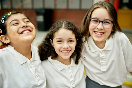 Waist-up view of 9 and 11 year old friends in matching uniforms standing with arms around each other, smiling and laughing while looking at camera.