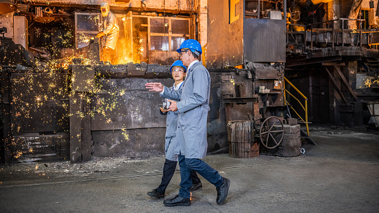 Male engineer explaining to his female colleague. Managers standing in foundry workshop. Dirty place and hot liquid metal in background.