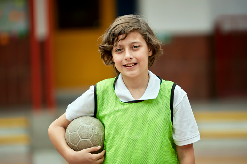 Waist-up view of elementary age Hispanic student wearing green scrimmage vest over polo shirt, holding ball under his arm, and smiling at camera.