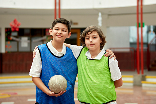 Waist-up view of elementary age classmates wearing blue and green scrimmage vests, standing with arms around each other, holding ball, and smiling at camera.