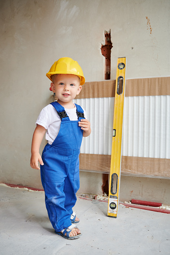 Full length of child plumber in work overalls standing by heating radiator with level ruler tool in apartment under renovation. Kid in safety construction helmet looking at camera and smiling.
