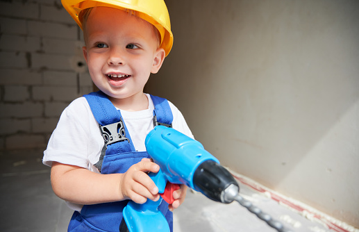 Cute baby boy construction worker looking aside and smiling while holding toy electric drill. Kid with power tool for repair works wearing safety helmet and work overalls. Home renovation concept.