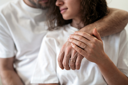Young heterosexual married couple with wedding rings on hands wearing white t-shirts hugging tenderly.