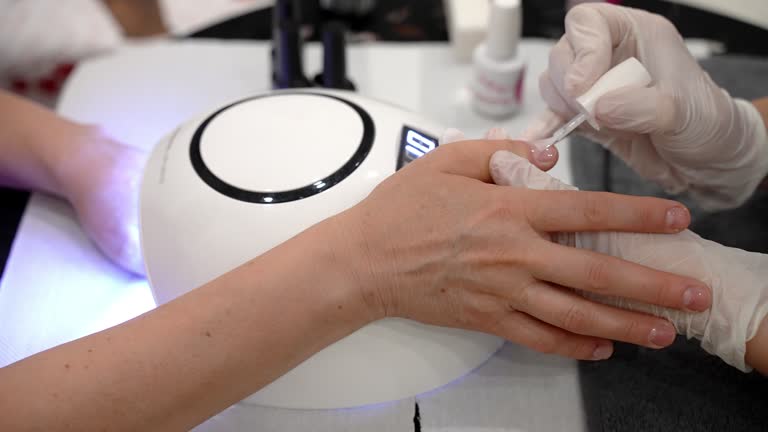 Beautician artist in disposable gloves applying transparent gel nail polish on woman's fingernail before drying under the ultraviolet lamp, modern trendy manicure procedures in beauty salon, toxic gel polish destructing nail plate and causing dermatologi