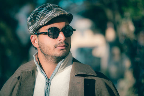 An Asian/Indian stylish young man stands outdoors and looks away on a sunny day holding a smartphone. He is in sunglasses, a cap, and a long coat.