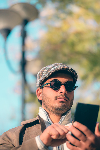 An Asian/Indian stylish young man uses a smartphone standing outdoors. He is in sunglasses, a cap, and a long coat.