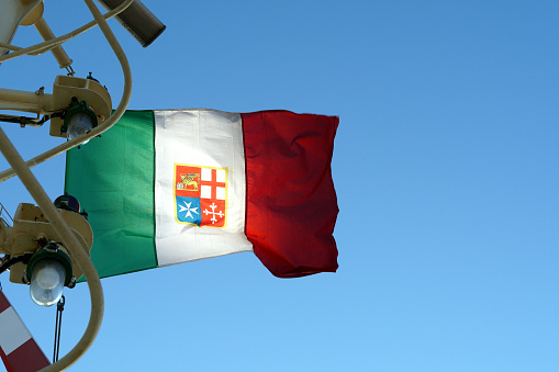 An Italian tricolour defaced with a variant of the arms of the Italian Navy and without a mural crow on the wind hanging on the navigational mast with lights and radar on merchant container vessel.