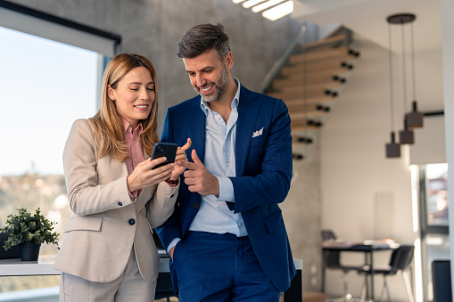 Smiling blond businesswoman holding smart phone having discussion with coworker male manager employee in office lobby, discussing project strategy, checking corporate plan on mobile app.