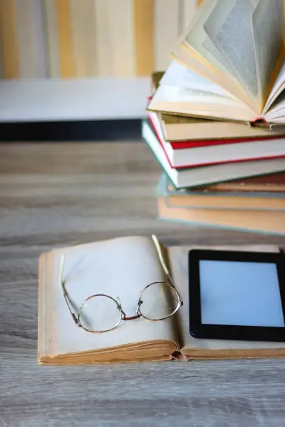 Stack of books on the table, reading glasses and e-reader on the table. Bookshelf in the background. Selective focus.