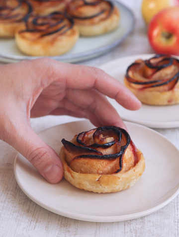 A baked mini caramelized apple rose tarts rolled in a rose form with puff pastry in a plate and hand going to grab it with its fresh fruit in background