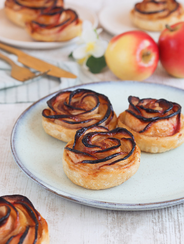 A baked mini caramelized apple rose tarts rolled in a rose form with puff pastry in a plate with knife and fork and its fresh fruit in background.