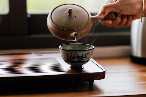 A cropped photo of an anonymous Asian female making traditional tea in a teapot and pouring it into the cup.