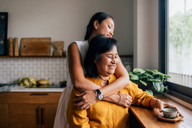 une belle femme heureuse serrant sa mère dans ses bras alors qu’elle est assise dans la cuisine et boit du thé - senior adult child holding grandparent photos et images de collection