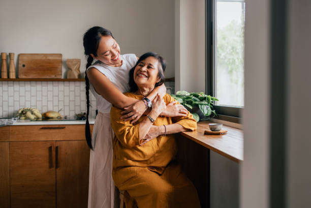 a happy beautiful woman hugging her mother while she is sitting in the kitchen and drinking tea - vrije tijd fotos stockfoto's en -beelden