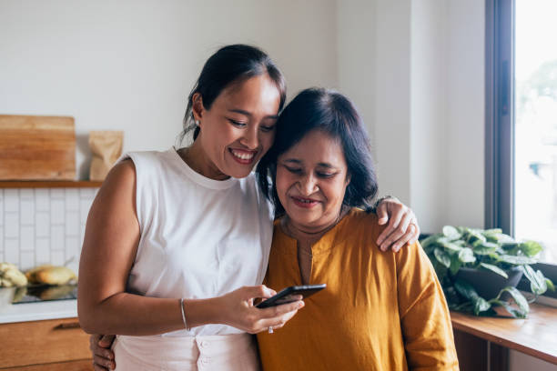 une belle femme heureuse regardant quelque chose sur son téléphone portable avec sa mère (elle l’embrasse) alors qu’ils sont debout dans la cuisine - two parent family indoors home interior domestic kitchen photos et images de collection