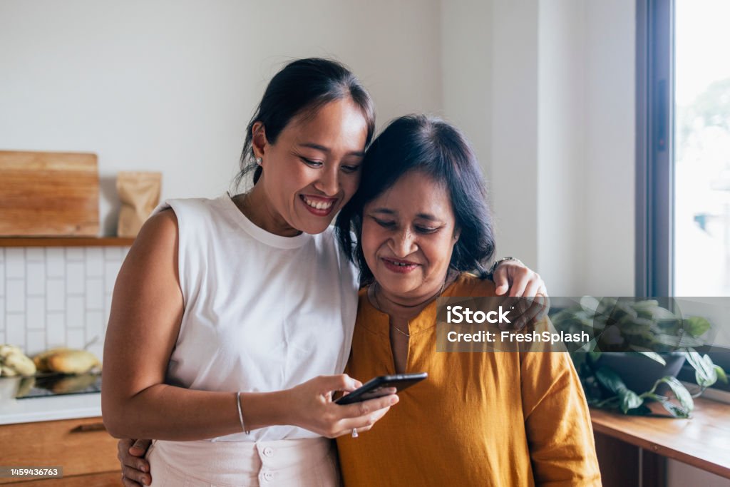 A Happy Beautiful Woman Watching Something On Her Mobile Phone With Her Mother (She Is Embracing Her) While They Are Standing In The Kitchen A smiling Asian female hugging her mother while they are reading something online using their black smartphone. Family Stock Photo