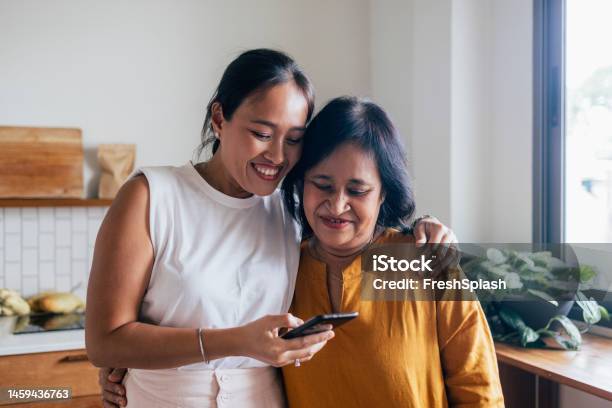 A Happy Beautiful Woman Watching Something On Her Mobile Phone With Her Mother While They Are Standing In The Kitchen Stock Photo - Download Image Now