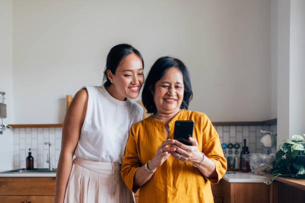 행복한 아름다운 노인 여자는 부엌에 서 있는 동안 예쁜 딸과 함께 휴대폰으로 무언가를 보고 있다 - family multi ethnic group asian ethnicity cheerful 뉴스 사진 이미지