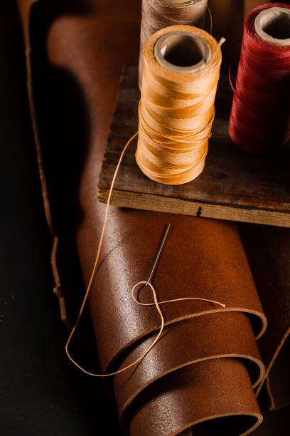 Close-up thread with needle on roll of brown genuine leather in craftsman workshop. Handmade genuine leather production in the factory. Close-up thread with needle on roll of brown genuine leather in craftsman workshop. Handmade genuine leather production in the factory sewing thread rolled up creation stock pictures, royalty-free photos & images