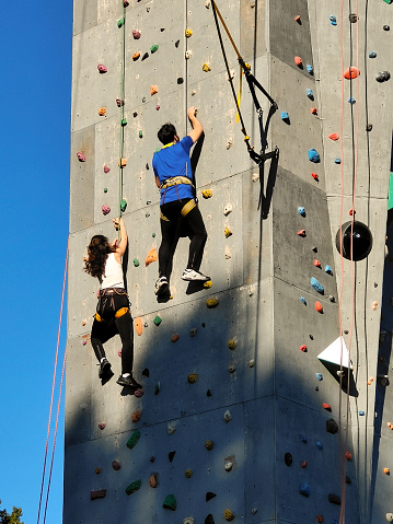 A boy and a girl compete on the wall to climb the hooks in special equipment. A wall on the street. View from below