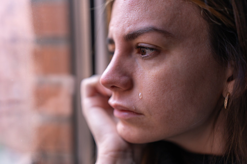 close-up of a depressed woman looking out the window, while a tear falls down her cheek. Depression and mental health concept