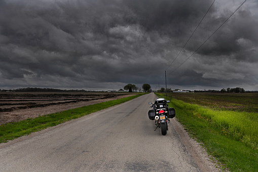 Port William, OH, USA – April 12, 2021.  A sport touring motorcycle is parked on the side of a remote rural road in farmland, about to ride into a huge storm moving across the region.  Dark threatening storm clouds loom overhead on this one lane road in western Ohio, heading towards an isolated farm complex.