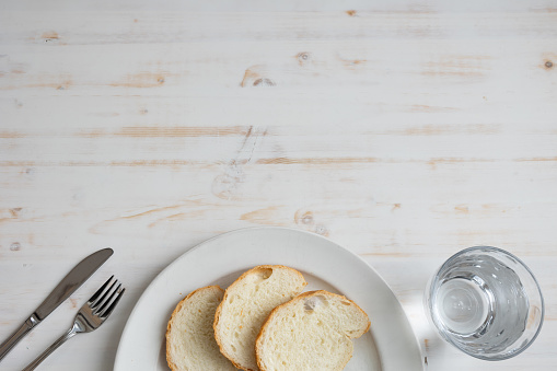 Border of a white plate with three slices of bread with a knife and fork and a glass of water on a white wood table with copy space shot from above