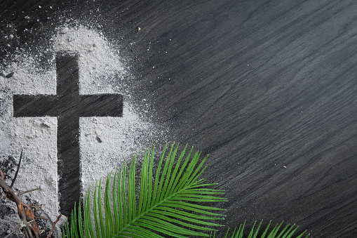 Christian lent background with cross of ashes, palm fronds and partial crown of thorns on a dark wood background with copy space