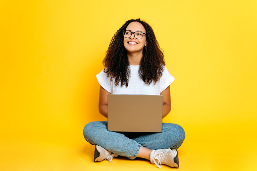 Full length photo of positive joyful creative young brazilian or hispanic woman with glasses, freelancer or designer, sit on isolated orange background with laptop, looks to the side, dreaming, smiles