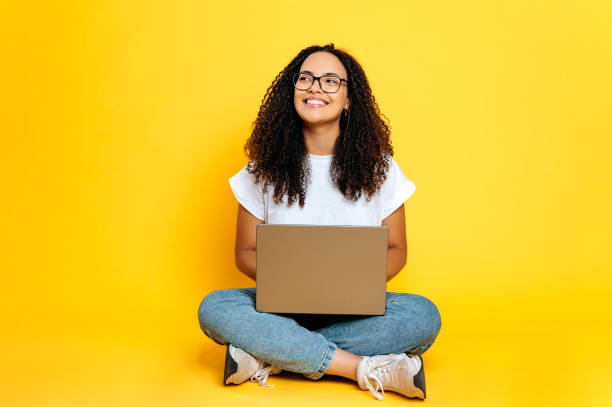 Full length photo of positive joyful creative young brazilian or hispanic woman with glasses, freelancer or designer, sit on isolated orange background with laptop, looks to the side, dreaming, smiles stock photo