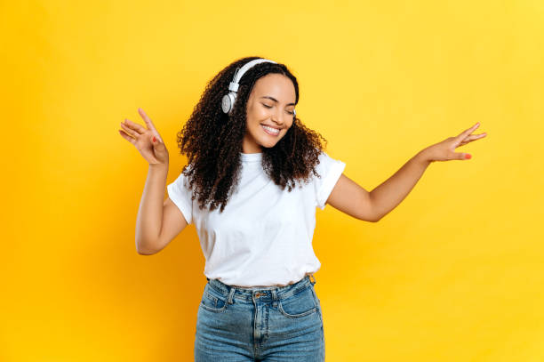 alegre encantadora joven morena de raza mixta, con ropa casual y elegante, con auriculares, escucha su música favorita, baila con los ojos cerrados, se relaja, se divierte sobre fondo naranja aislado, sonríe - music women disco joy fotografías e imágenes de stock