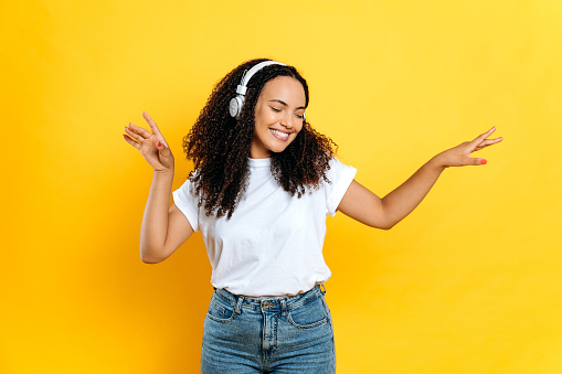 Cheerful lovely young brunette mixed race woman, in casual stylish wear, with headphones, listens her favorite music, dancing with eyes closed, relaxing, having fun on isolated orange background,smile