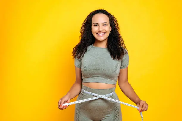 Photo of Positive lovely sporty mixed race young woman, in sports outfit, stand on isolated orange background, holding centimeter tape and measuring her waist, happy with results, lose weight, smiles at camera