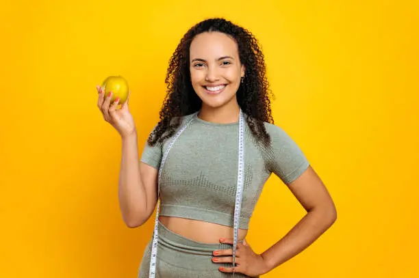 Photo of Beautiful confident brazilian or hispanic curly haired sporty woman, in sports outfit, holds fresh apple, standing on isolated orange background, looking at camera with happy smiling facial expression