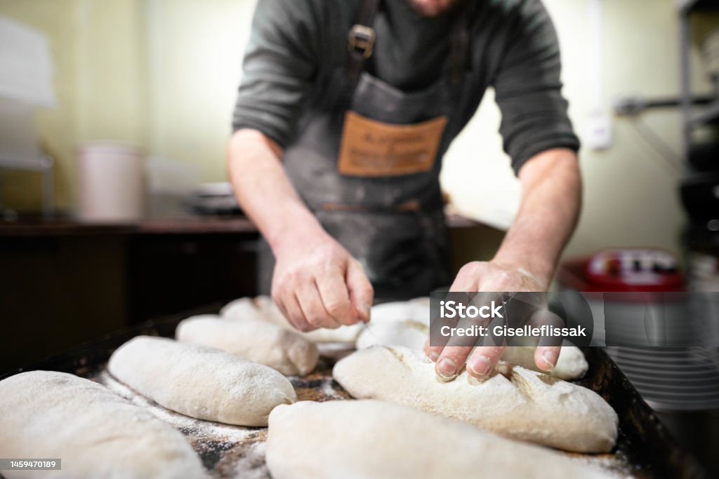 Baker scoring loaves of bread before baking them in an oven Close-up of a baker scoring loaves of bread on a tray with a knife before putting them in an oven in a commercial kitchen Bakery Stock Photo