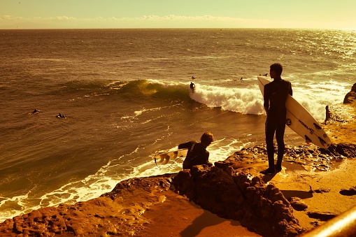 Santa Cruz, United States - January 27 , 2013 : Looking at the sunset of santa barbara bay while this surfer is standing on a rock with his board in his arms.