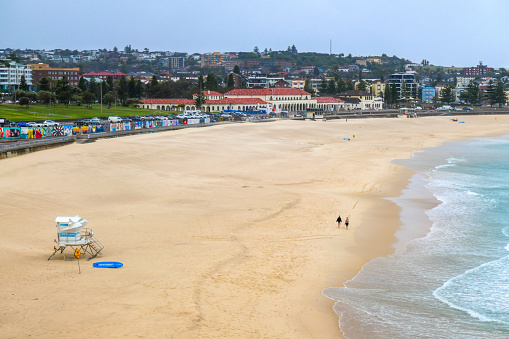 Two people walking along the water's edge at Bondi Beach, Sydney on an overcast, windy and rainy day on 5 January 2023.  This image was taken in the afternoon from the cliffs at the southern end of the beach looking north.