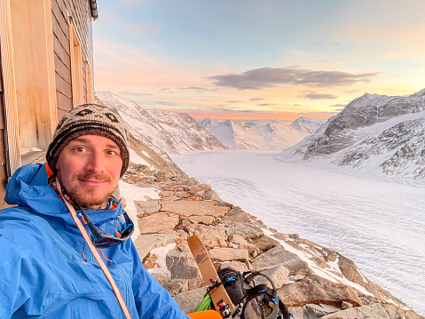 Male mountaineer climbing up mountain in winter, he takes selfie near mountain hut