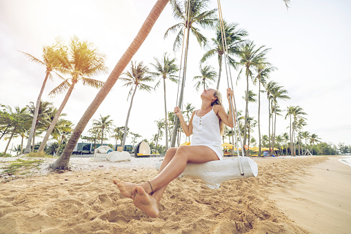 Simple life concept, happy woman on swing at the beach enjoying her life under the palm trees. Tropical vacation destination.