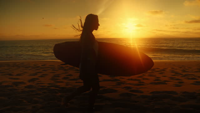Young woman with long hair is walking on the waterfront during sunset,carrying a surfboard underneath her arm